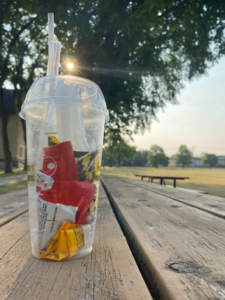 Clear plastic to-go cup filled with plastic waste, placed on a picnic table in front of trees,
