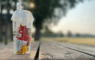 A clear to-go cup, filled with plastic wrappers and waste, sitting on top of a picnic table. Blurry trees in the background.