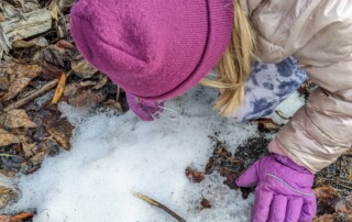 girl in pink coat and purple hat and gloves kneeling in snow looking through a magnifying glass