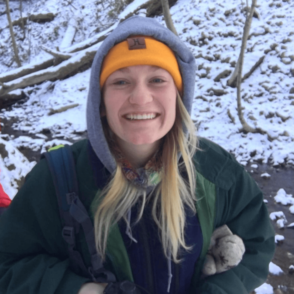 Young woman smiling at the camera, during a winter hike. The background is a snowy forest.