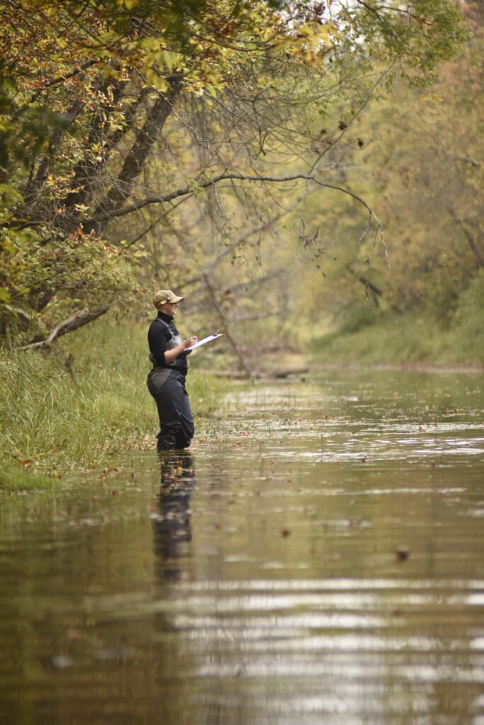 A person standing knee-deep in a river holding a clipboard